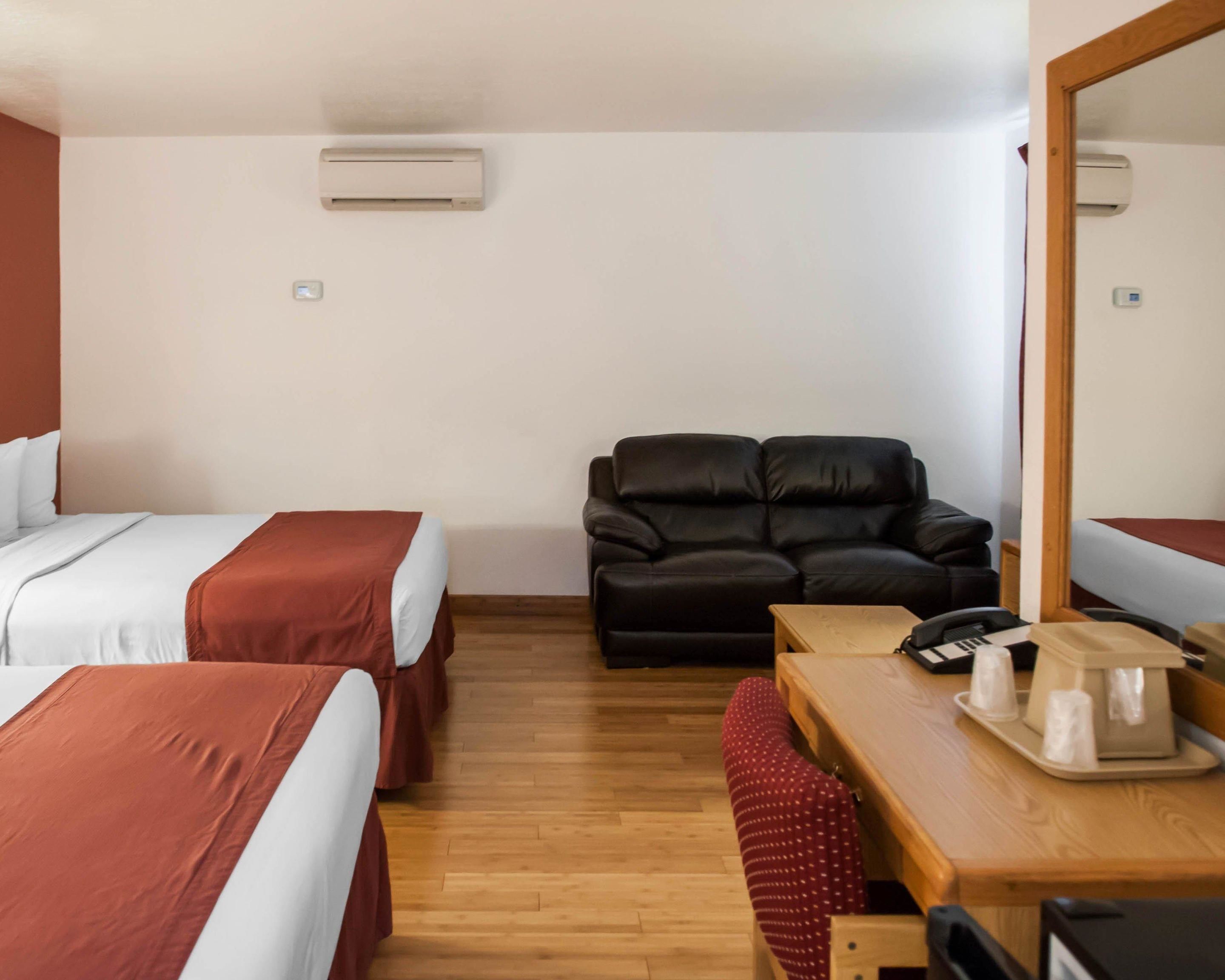 A hotel room with two beds, a sofa, a wooden table with a phone and a coffee maker, and an air conditioning unit on the wall.