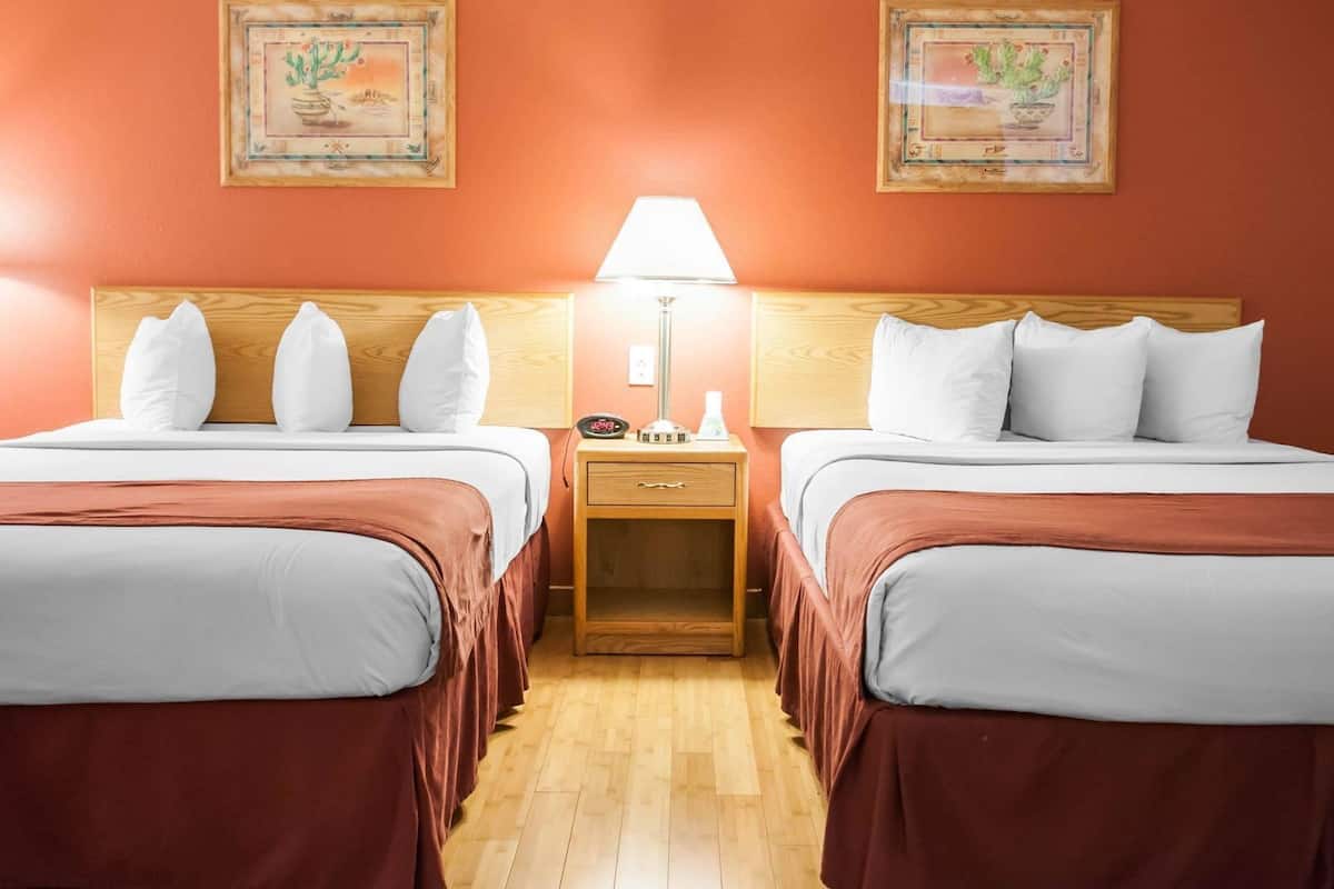 Quarto de hotel com duas camas, cabeceiras de madeira, uma mesa de cabeceira com abajur e duas fotos emolduradas na parede.