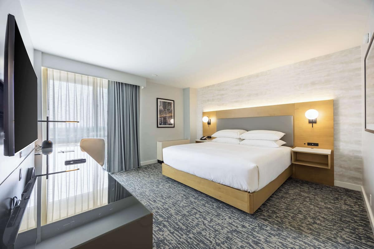 Habitación de hotel con una cama grande, un escritorio con silla, televisor y un ventanal con cortinas.