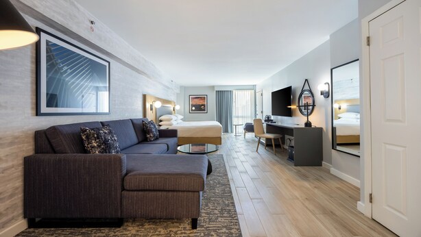 Ein modernes Hotelzimmer mit einer Eckcouch, einem Bett, einem Schreibtisch und einem Fernseher.