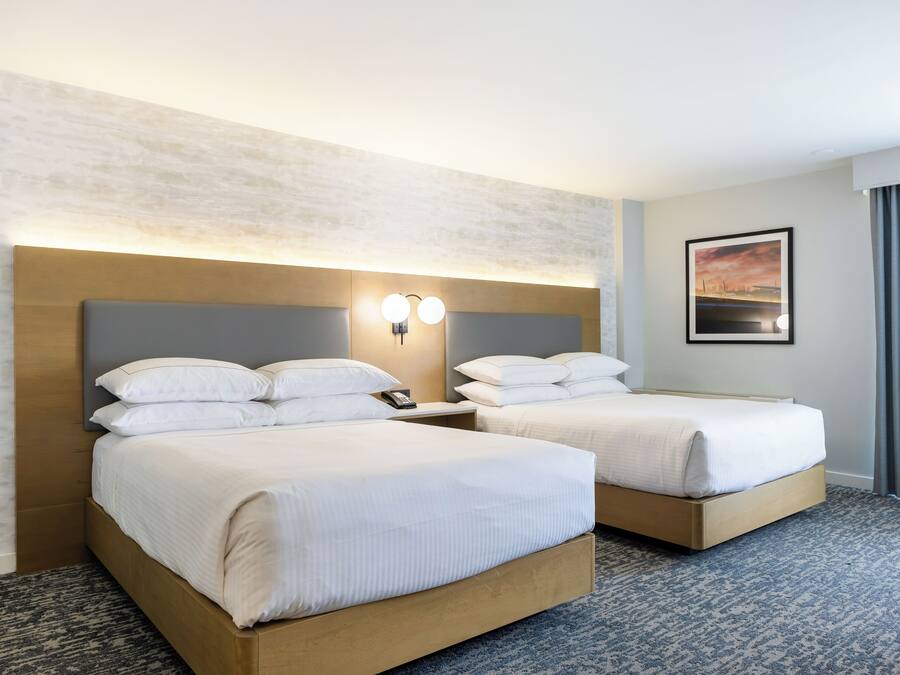 A hotel room with two beds, a nightstand with a phone, a wall-mounted light fixture, and a framed picture on the wall.