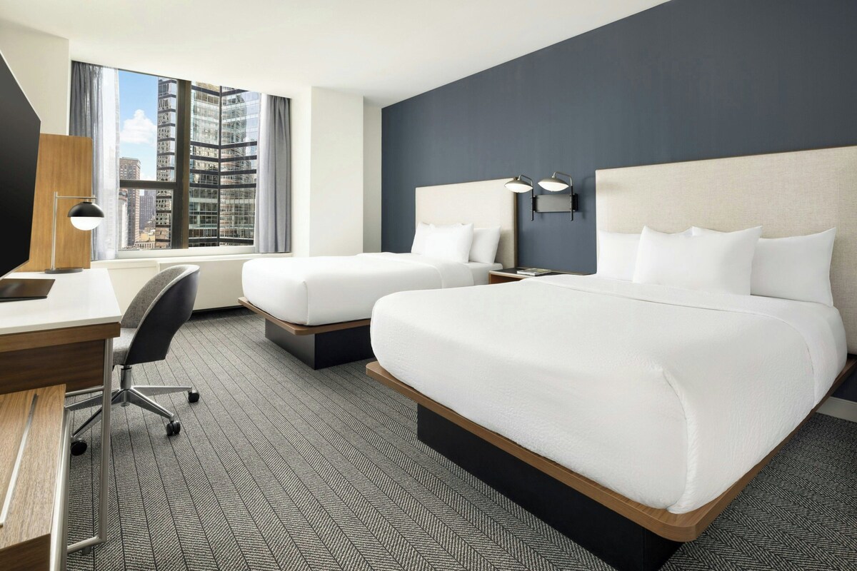 Quarto de hotel com duas camas, uma mesa, uma cadeira, um abajur e vista para a paisagem urbana.