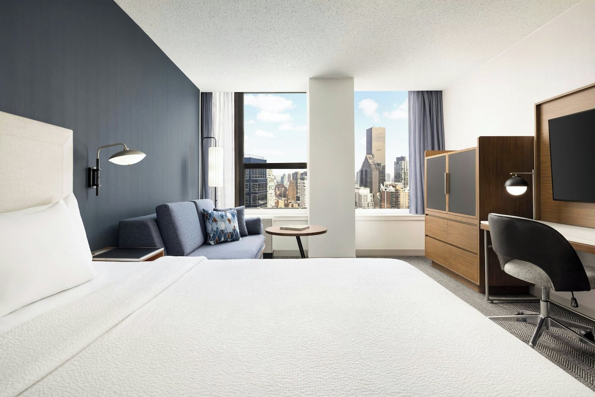 A hotel room with a large bed, a sofa, a desk, a chair, a TV, and a view of the city.