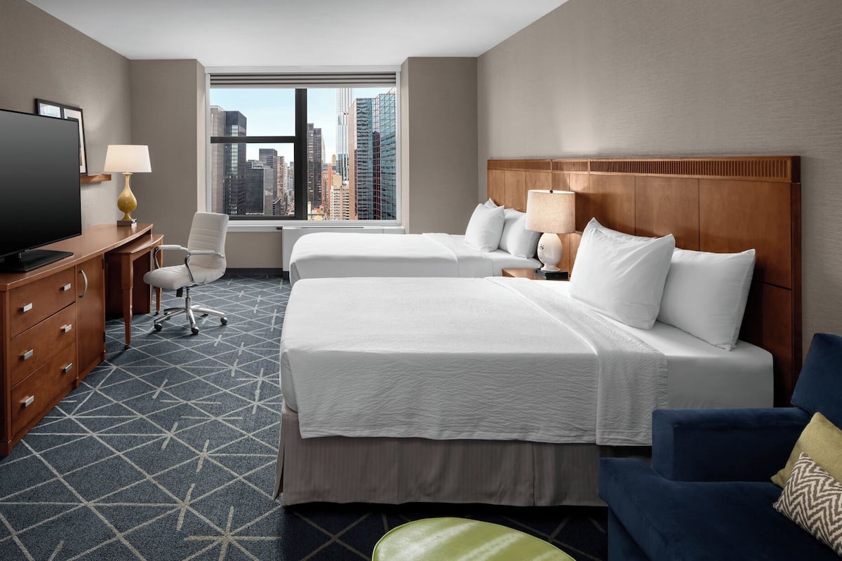 A hotel room with two beds, a desk, a chair, a TV, and a view of the city skyline.