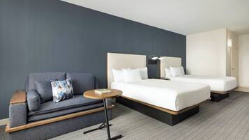 A hotel room with a grey sofa, a small round table, two beds with white bedding, and a grey herringbone-patterned carpet.