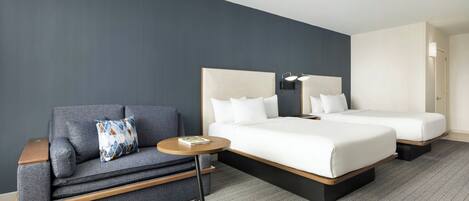 A hotel room with a grey sofa, a small round table, two beds with white bedding, and a grey herringbone-patterned carpet.