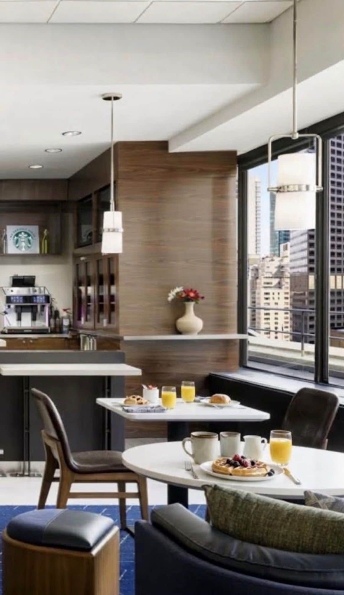 A modern hotel room with a kitchenette, dining area, and a large window with a city view.