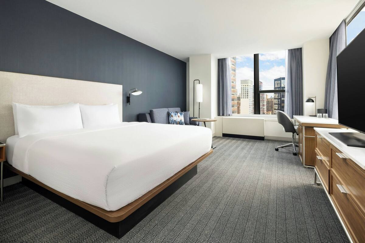 A hotel room with a large bed, a desk, a chair, a TV, and a window with a city view.
