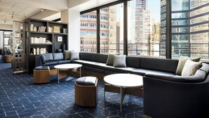 Un espace de détente moderne comprenant un canapé, des poufs et des tables, de grandes fenêtres offrant une vue sur la ville, ainsi qu’une bibliothèque.