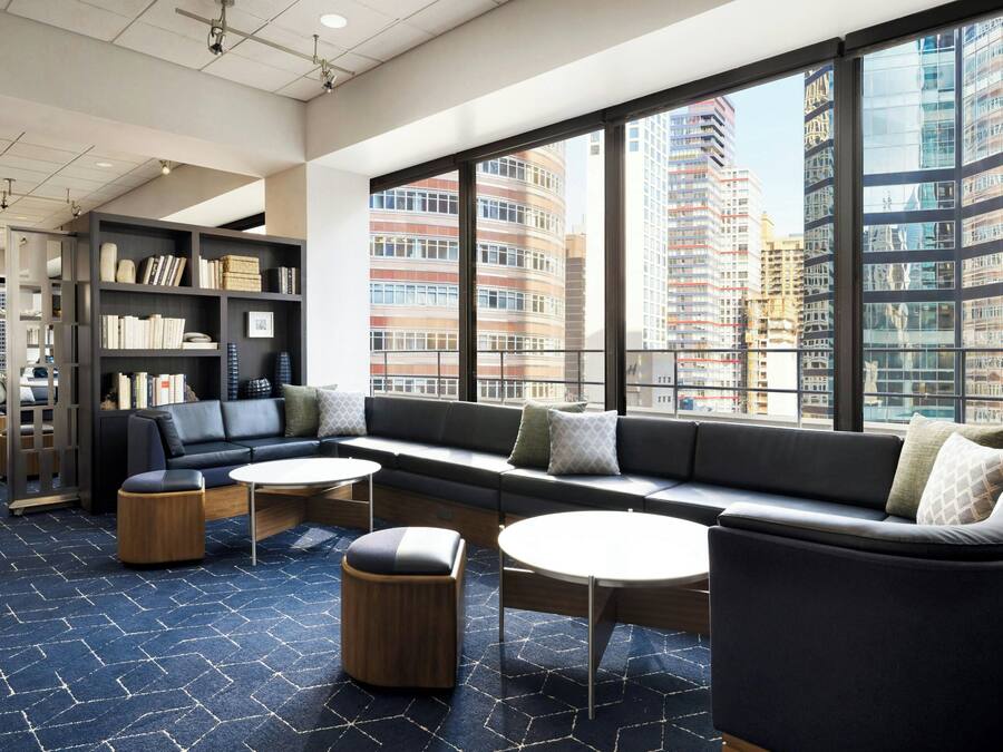 A modern lounge area with a sofa, ottomans, and tables, large windows with a city view, and a bookshelf.