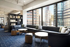 Een moderne zithoek met een bank, poefs en tafels, grote ramen met uitzicht op de stad, en een boekenkast.