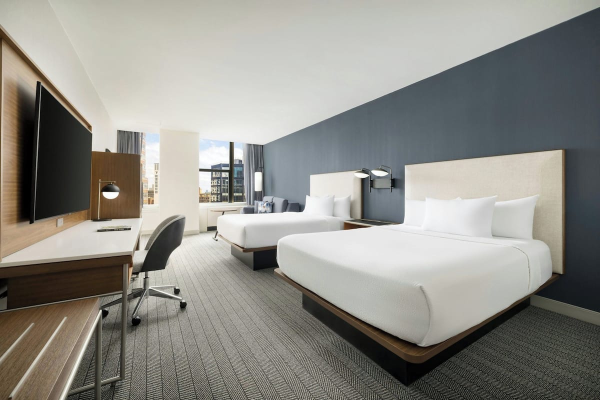 A hotel room with two beds, a desk, a TV, and a view of the city.
