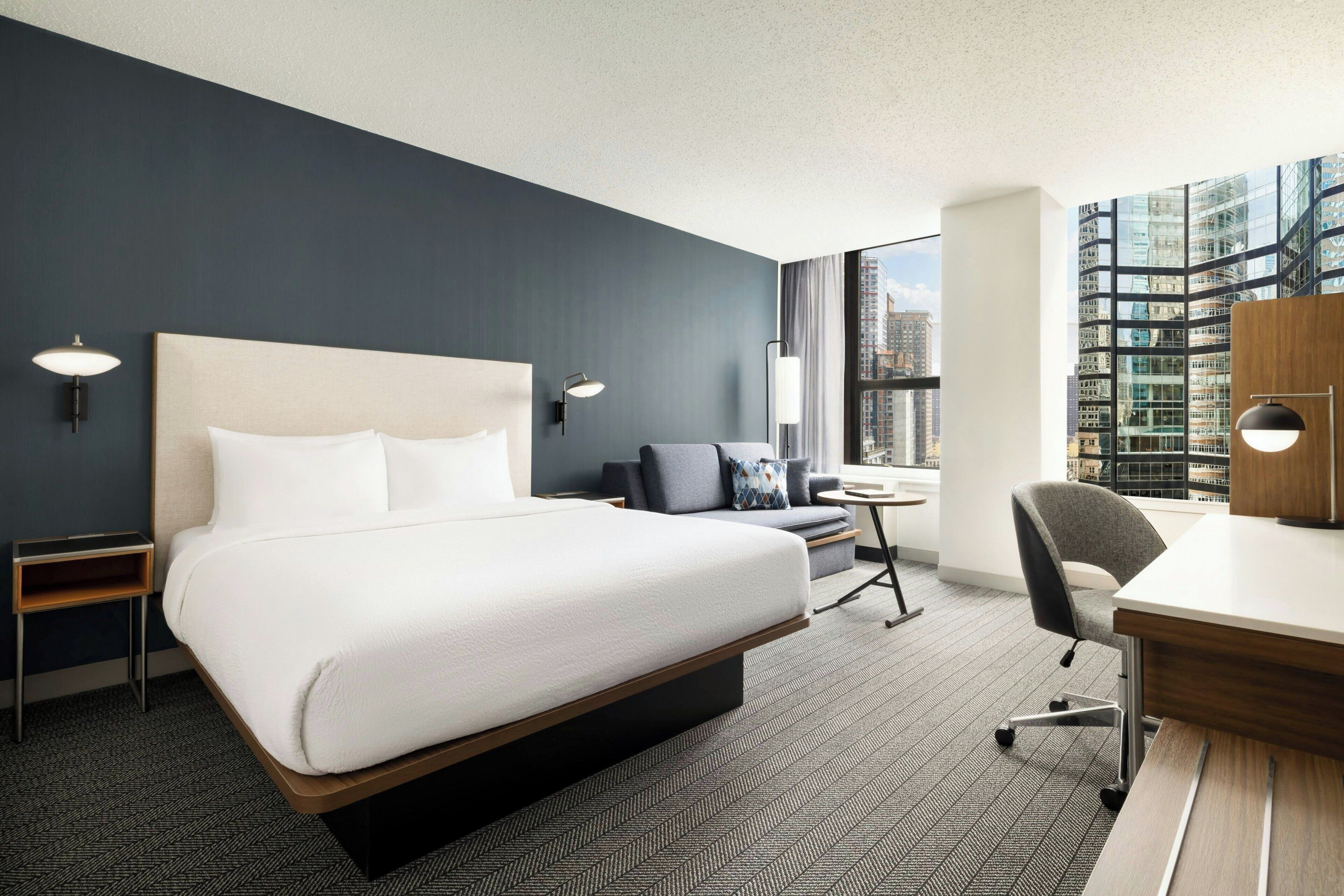 Une chambre d’hôtel moderne dotée d’un grand lit, d’un bureau, d’une chaise et offrant une vue sur le paysage urbain grâce à de grandes fenêtres.