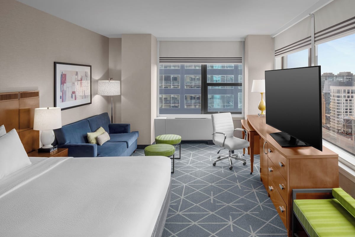 A hotel room with a large bed, a blue sofa, a desk with a TV, and a window with a city view.