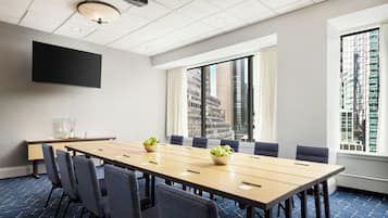 A conference room with a long table, chairs, a projector, and a large window with a city view.