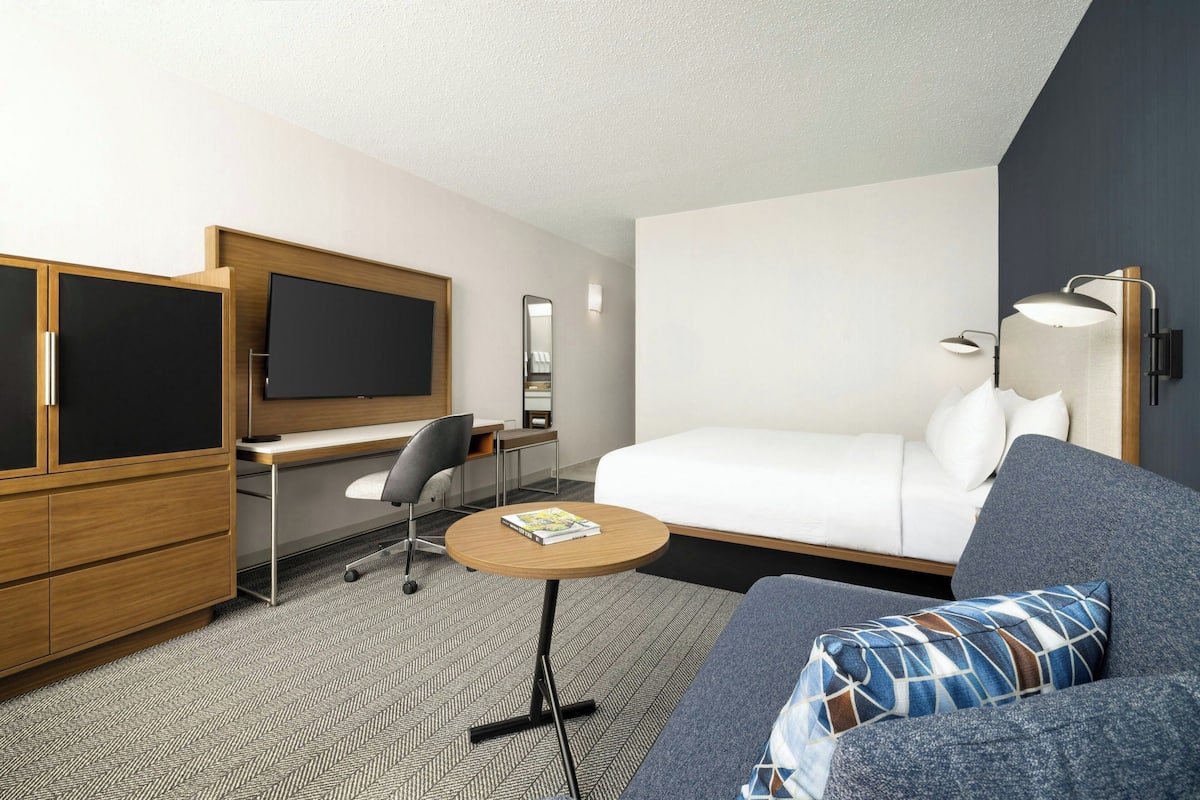 A modern hotel room with a bed, a desk, a chair, a round table, a TV, and a lamp.