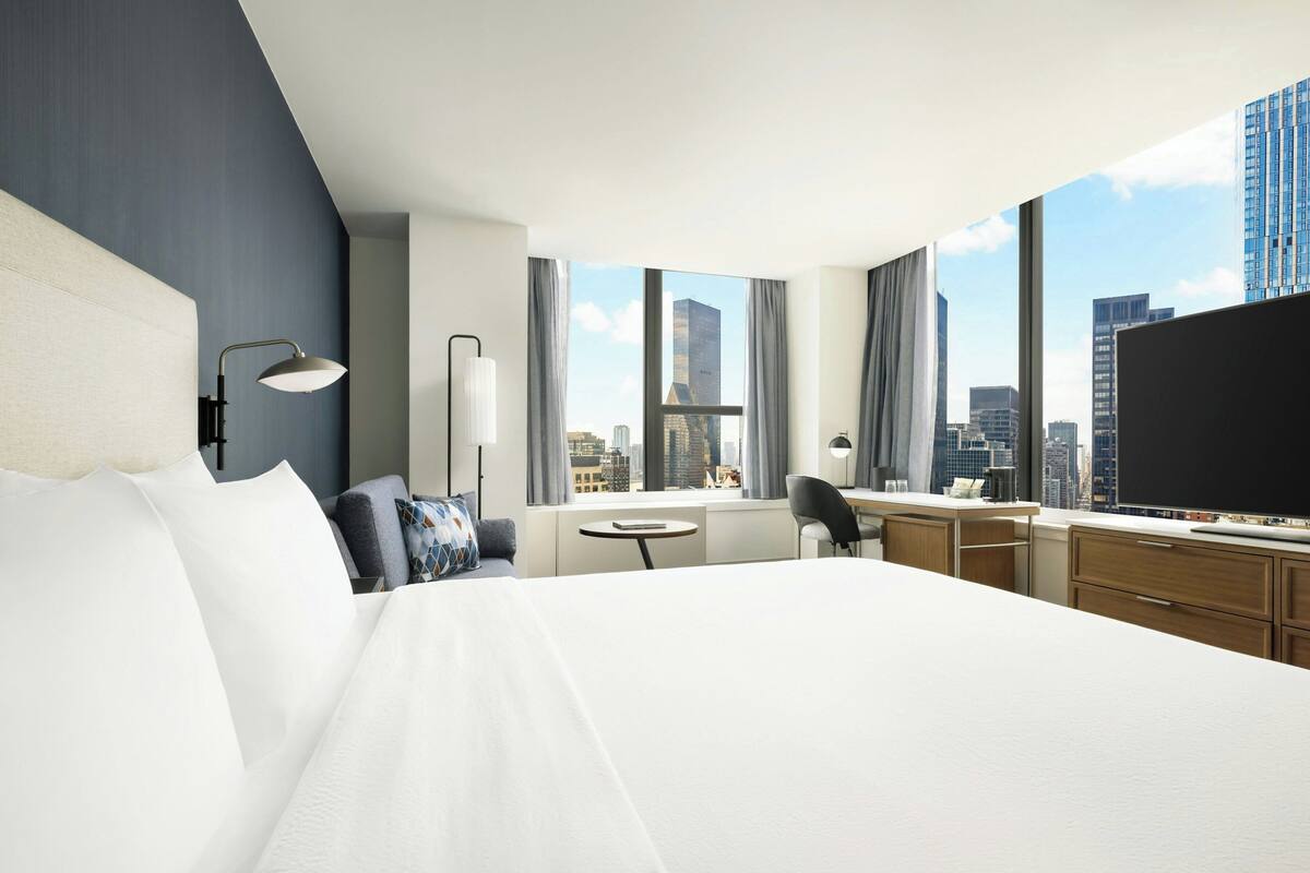 A hotel room with a large bed, a desk, a chair, a TV, and a view of the cityscape.