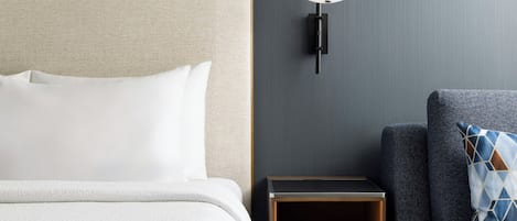 A neatly made bed with white linens, a wall-mounted lamp, a small wooden nightstand with a book, and a grey sofa with a patterned pillow.