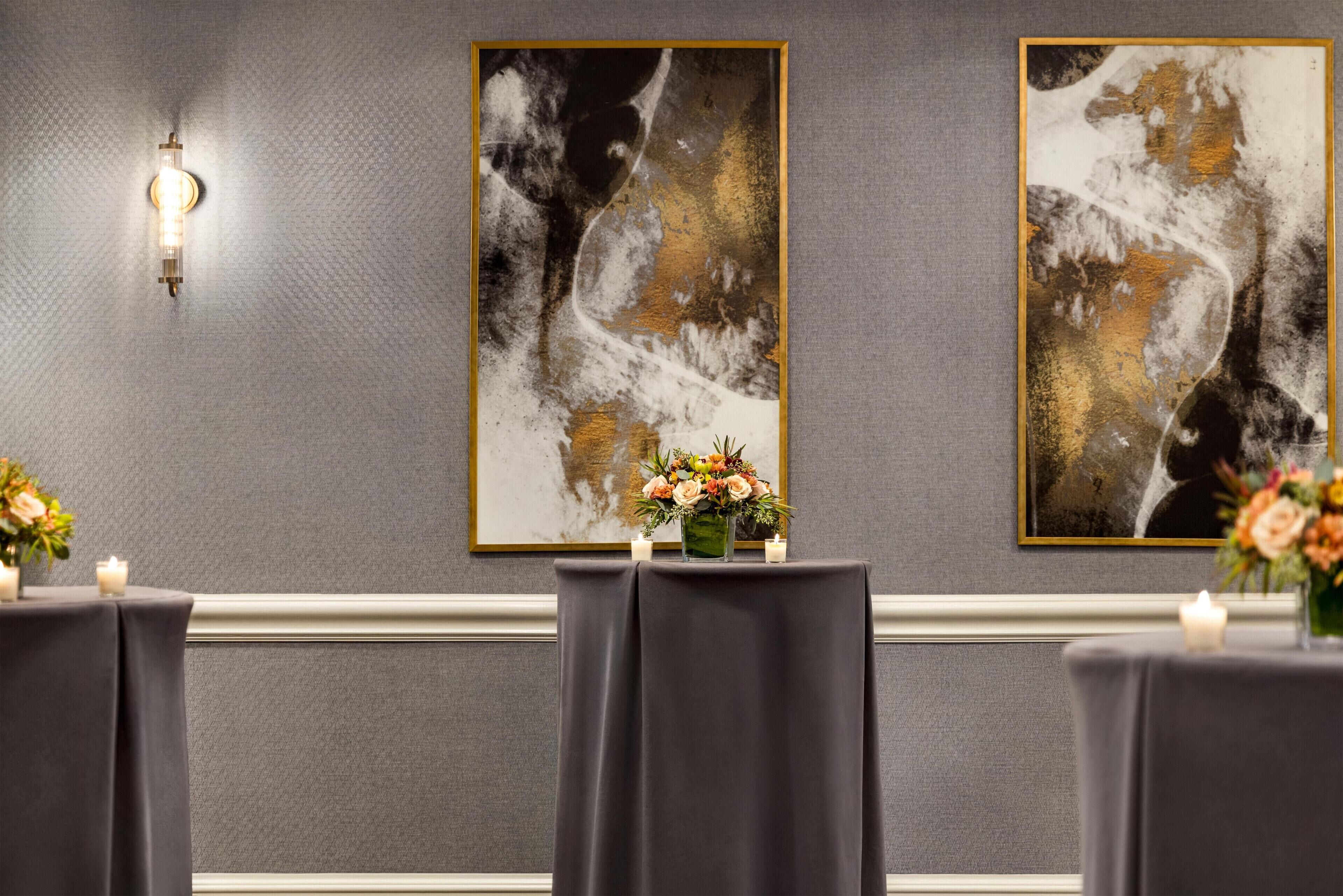 A room with a grey textured wall, two framed abstract paintings, a table with a dark tablecloth, and candles.