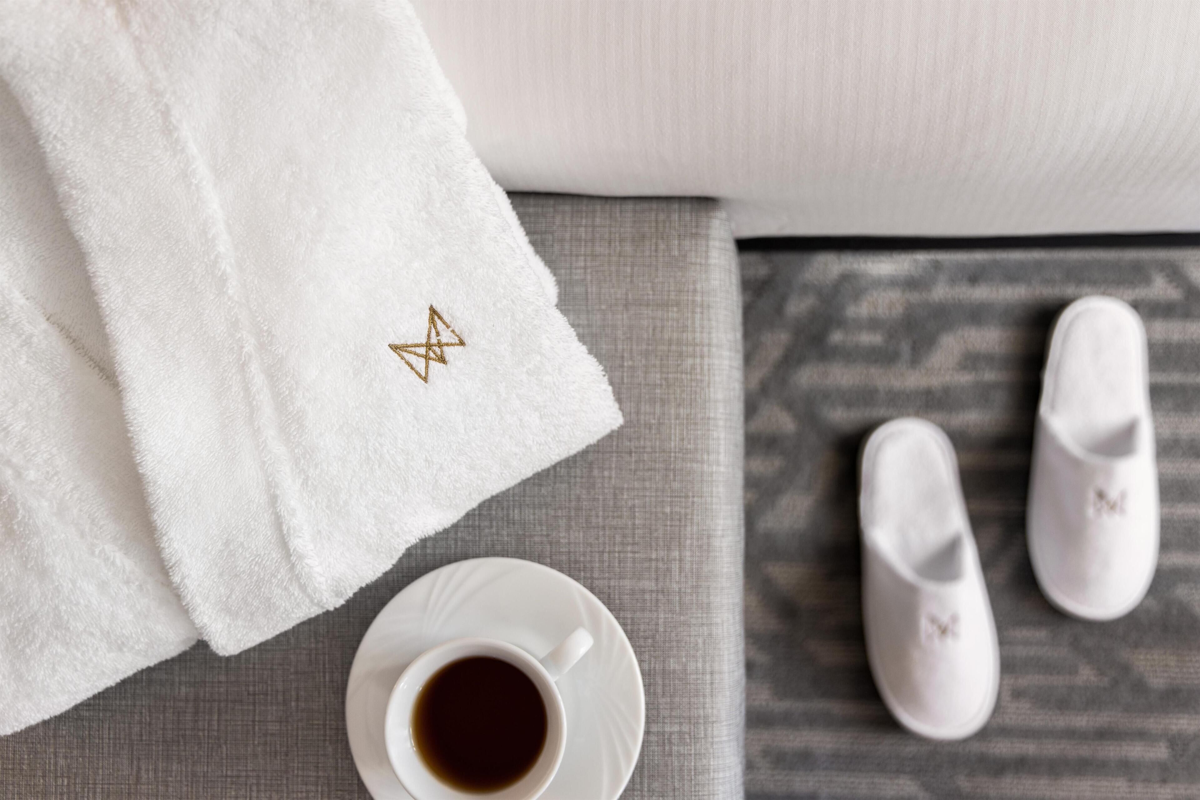 A white towel with a gold embroidered star, a cup of coffee, and a pair of white slippers on a grey textured surface.