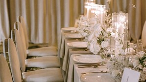 Una mesa de banquete dispuesta con platos blancos, cubertería dorada y centros de mesa florales.