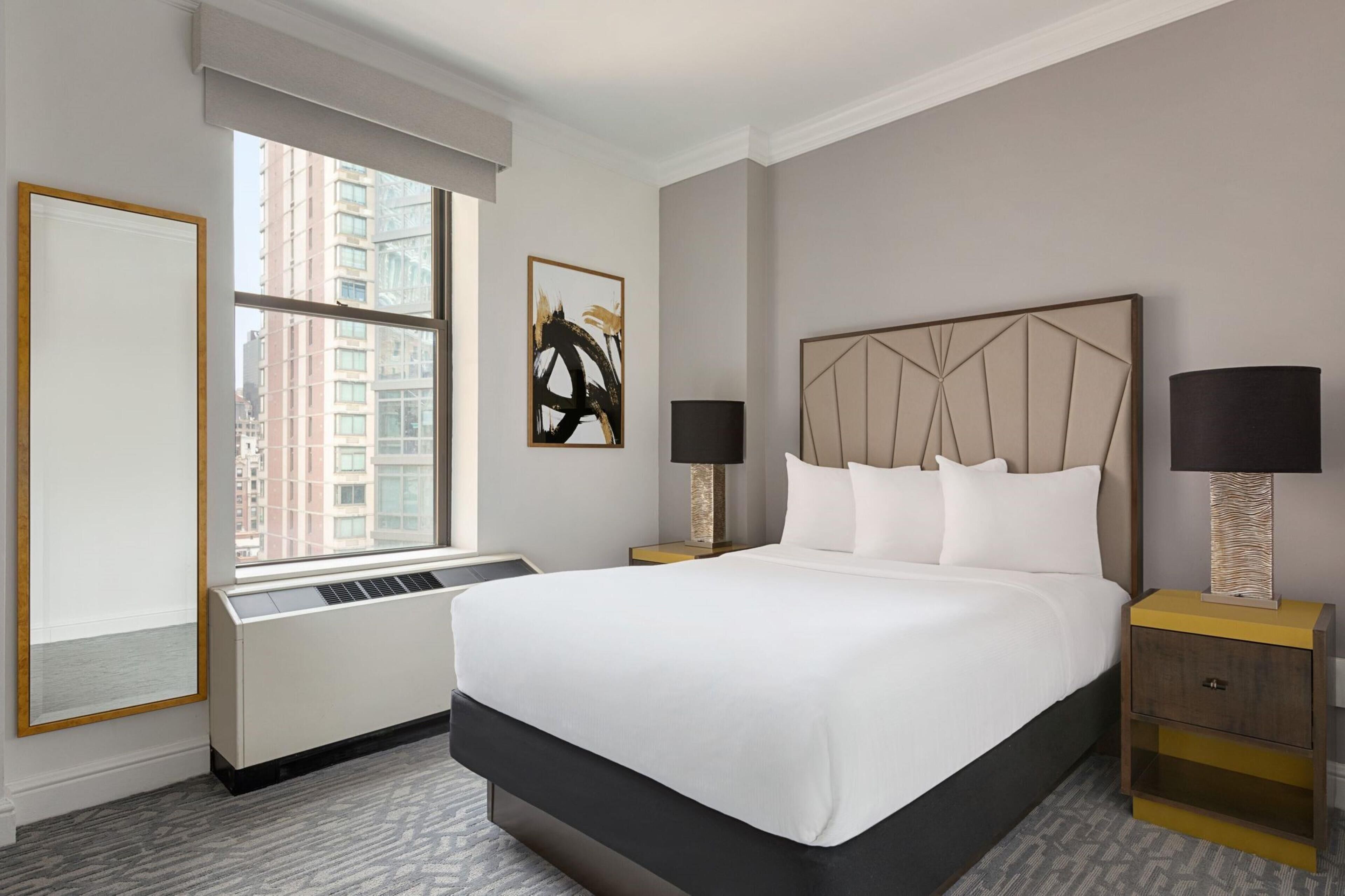 A hotel room with a large bed, two bedside tables, a window with a view of buildings, and a radiator.