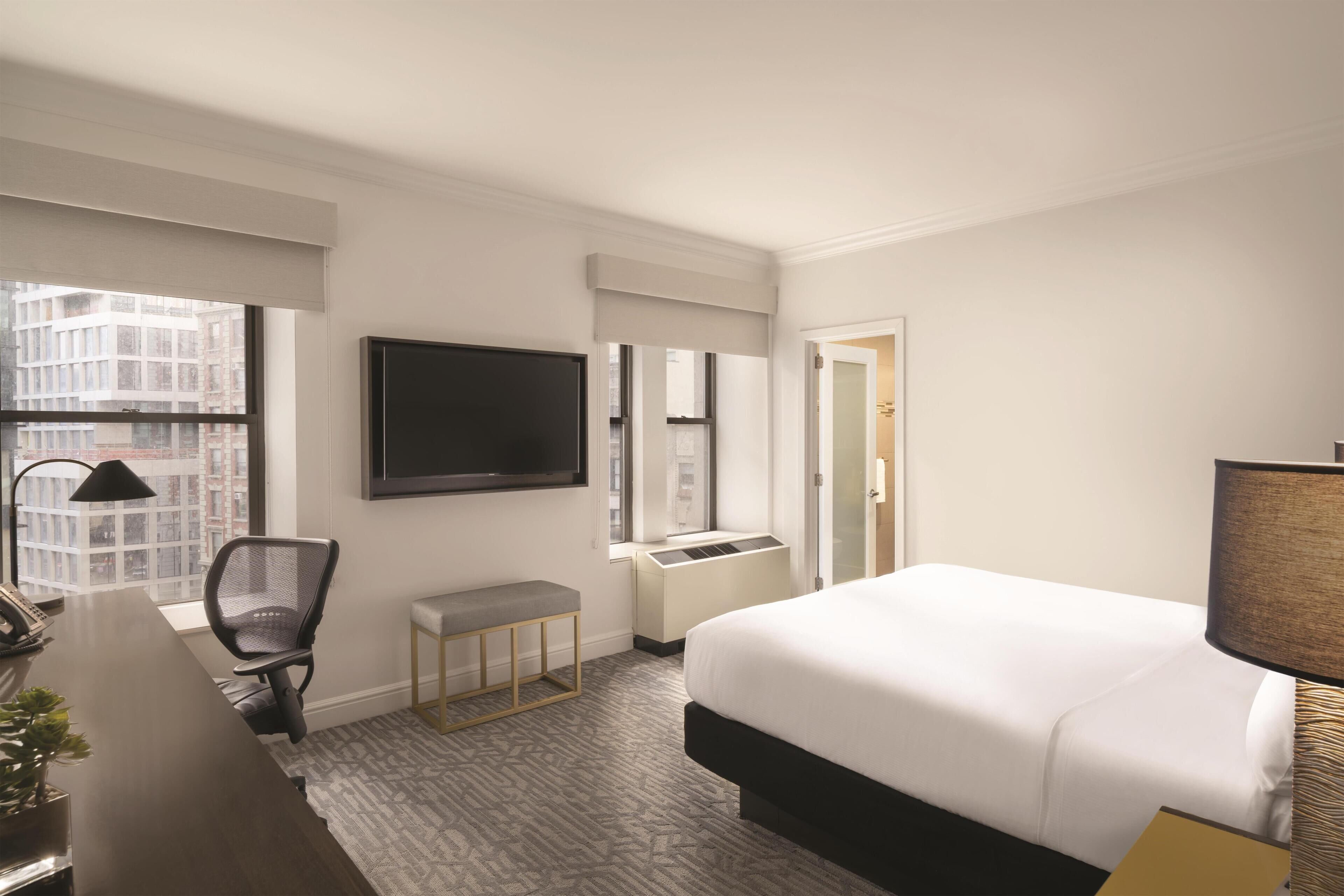 A hotel room with a large bed, a desk with a chair, a television, and a window with a view of buildings.