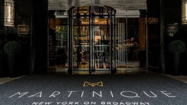 La entrada al hotel Martinique New York en Broadway, con una puerta giratoria, puertas de vidrio y un letrero con el nombre del hotel.