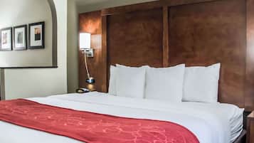 A hotel room with a large bed, a wooden headboard, a red throw blanket, and a nightstand with a lamp.