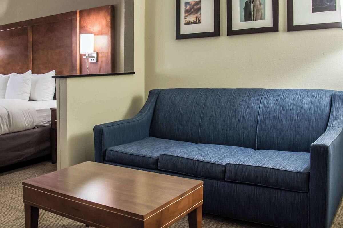 Une chambre d’hôtel avec un canapé bleu, une table basse en bois et un lit recouvert de draps blancs. Trois tableaux encadrés ornent le mur.