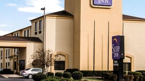 A Sleep Inn hotel with a beige facade, a prominent sign, and a parking lot with cars and trimmed bushes.