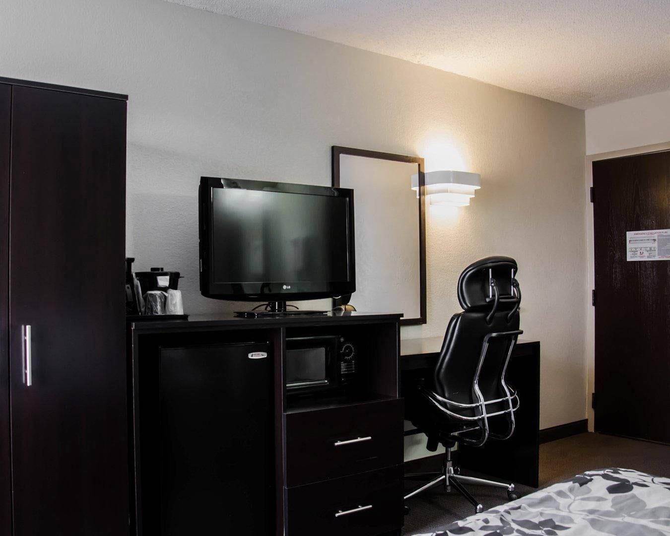 A hotel room with a dark wooden desk, a flat-screen TV, a black office chair, and a wall-mounted light fixture.