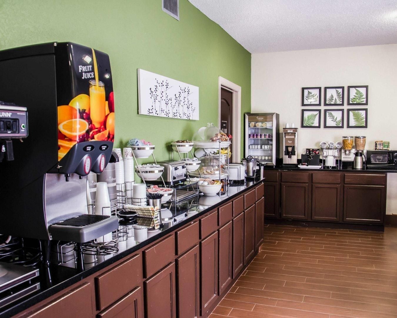 A self-service breakfast bar with a fruit juice dispenser, coffee machines, and a variety of food items.