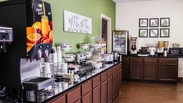 A self-service breakfast bar with a fruit juice dispenser, coffee machines, and a variety of food items.