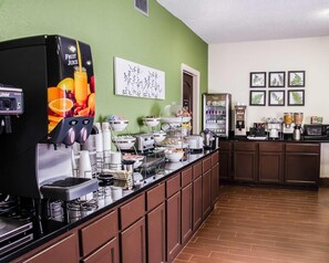 A self-service breakfast bar with a fruit juice dispenser, coffee machines, and a variety of food items.