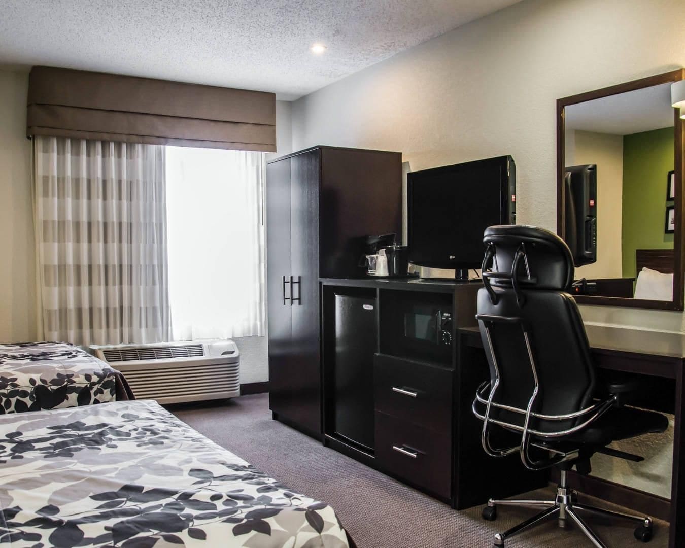 A hotel room with a bed, a desk with a computer, a chair, a mirror, and a window with curtains.