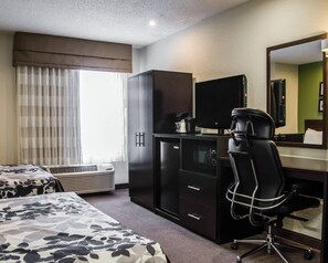 A hotel room with a bed, a desk with a computer, a chair, a mirror, and a window with curtains.