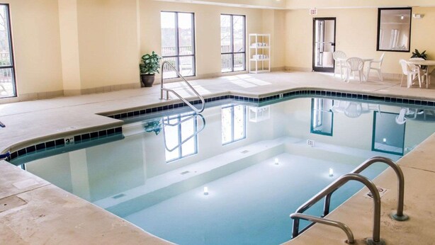 An indoor swimming pool with clear glass walls, a tiled deck, and a seating area with white chairs.