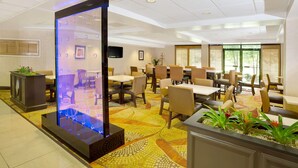 Un espace de restauration d’hôtel comprenant des tables, des chaises, un élément décoratif aquatique et une grande fenêtre laissant entrer la lumière naturelle.