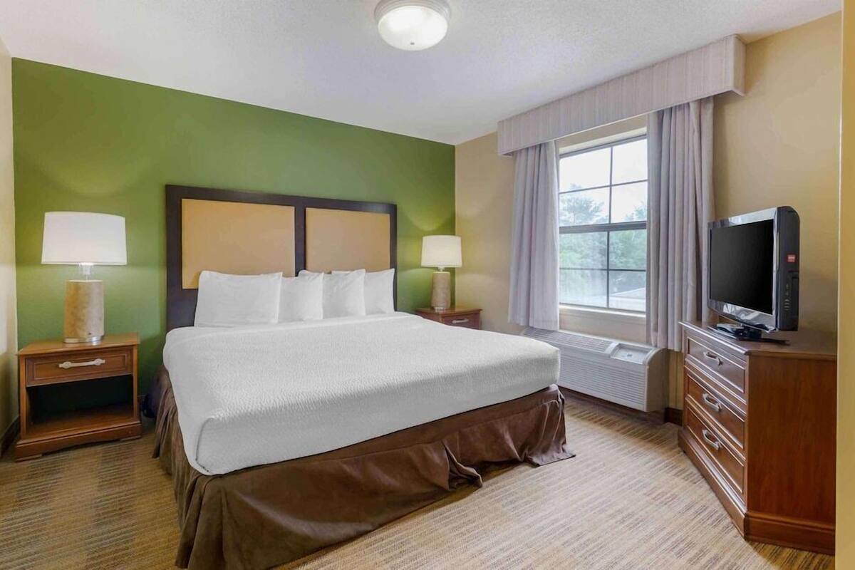 A hotel room with a large bed, two bedside tables with lamps, a television, and a window with curtains.