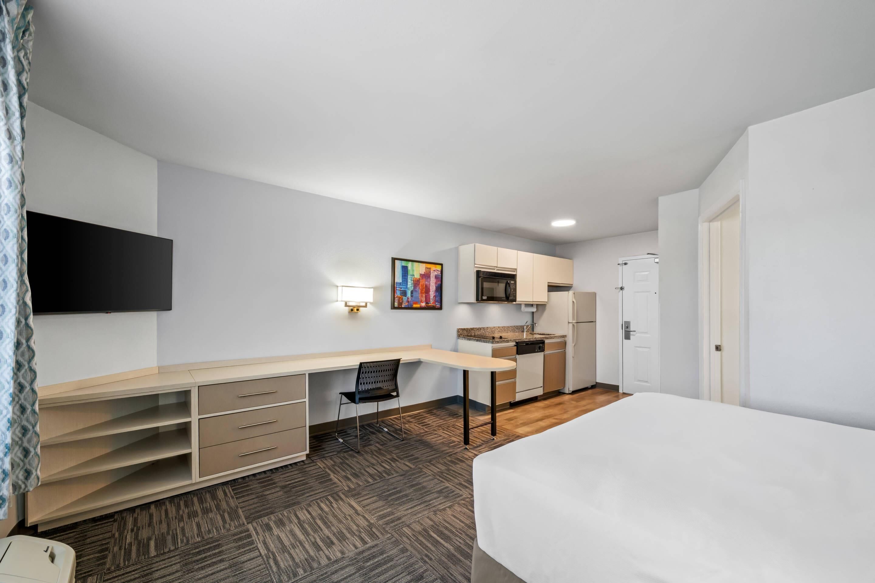 A compact hotel room with a bed, a desk, a chair, a kitchenette, and a TV mounted on the wall.