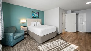A hotel room with a bed, a blue armchair, a nightstand with a lamp, and a painting on the wall. - MainStay Suites Greensboro (Greensboro)