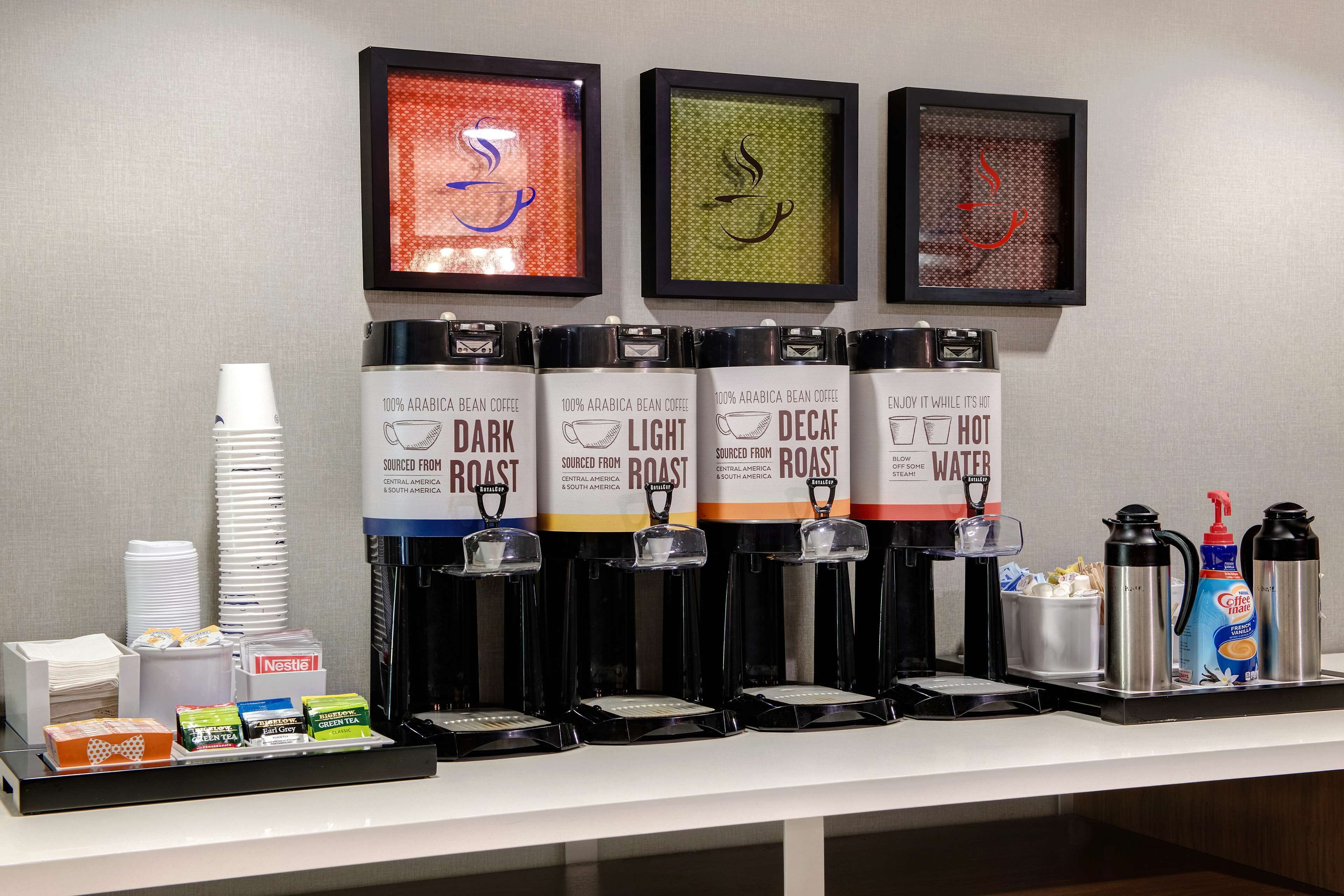A coffee station with multiple dispensers labeled 'Dark Roast,' 'Light Roast,' 'Decaf,' and 'Hot Water.' There are disposable cups, a coffee maker, and a bottle of coffee creamer.