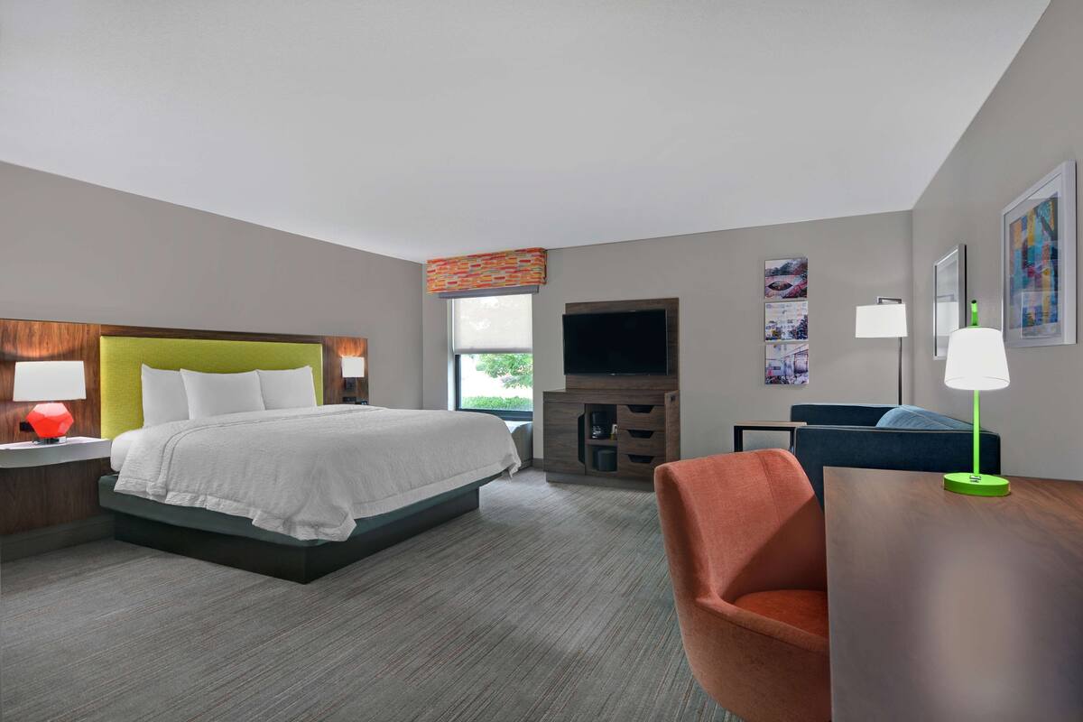 A hotel room with a bed, a desk, a chair, a TV, and artwork on the walls.