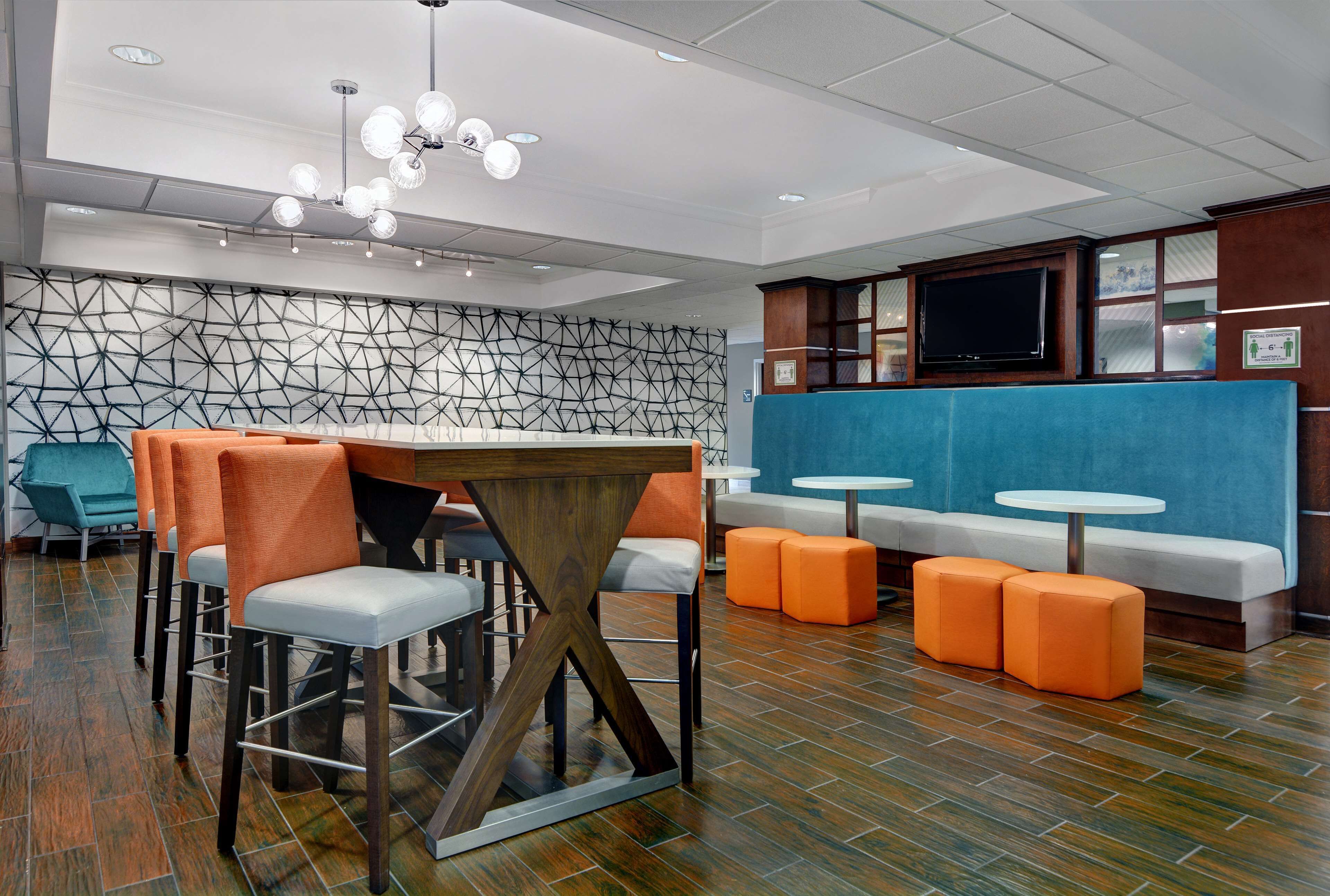 Un interior moderno con un muro de diseño geométrico, una mesa de madera con sillas, asientos naranja y una televisión.