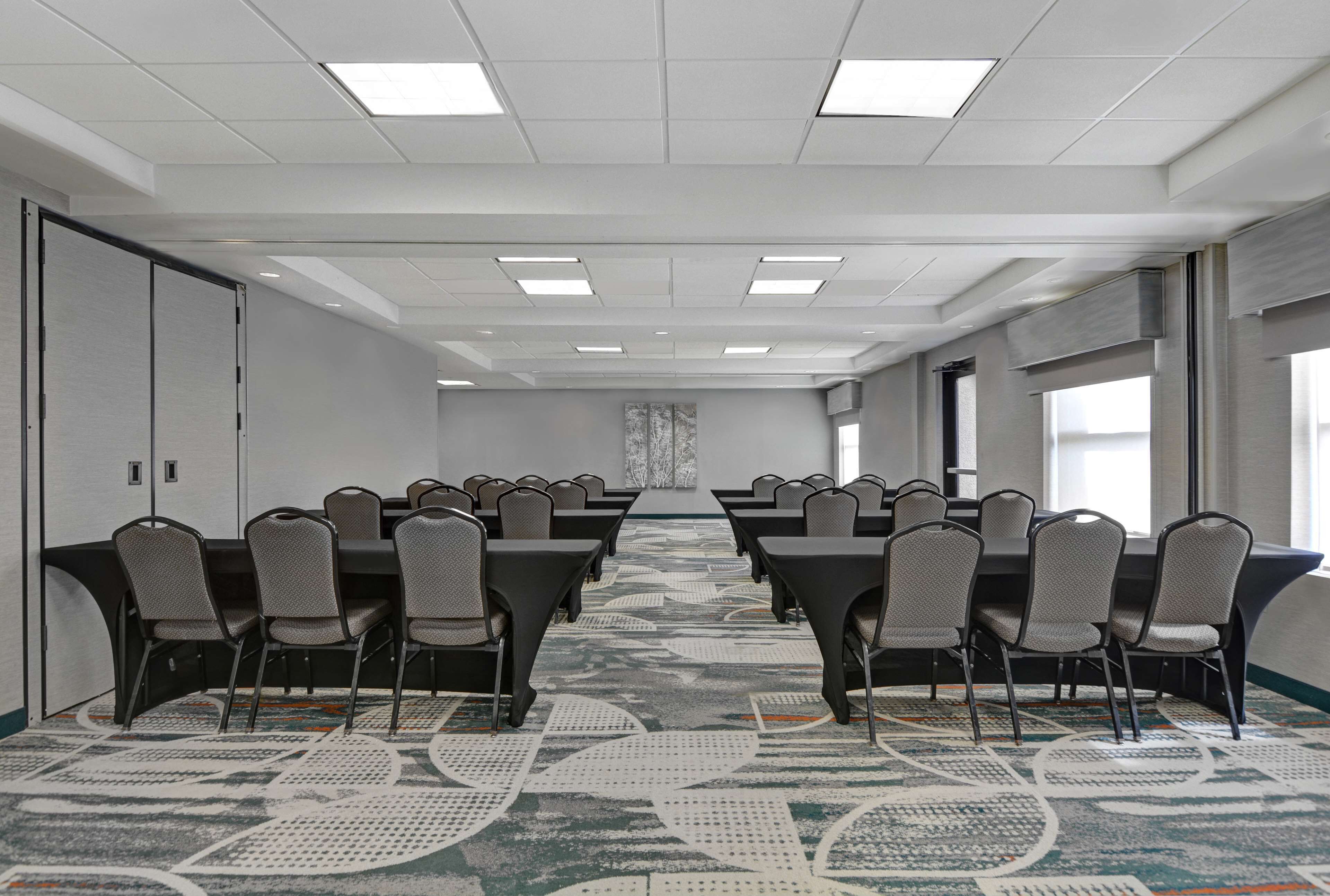 A conference room with rows of tables and chairs, a patterned carpet, and a large window on the right.