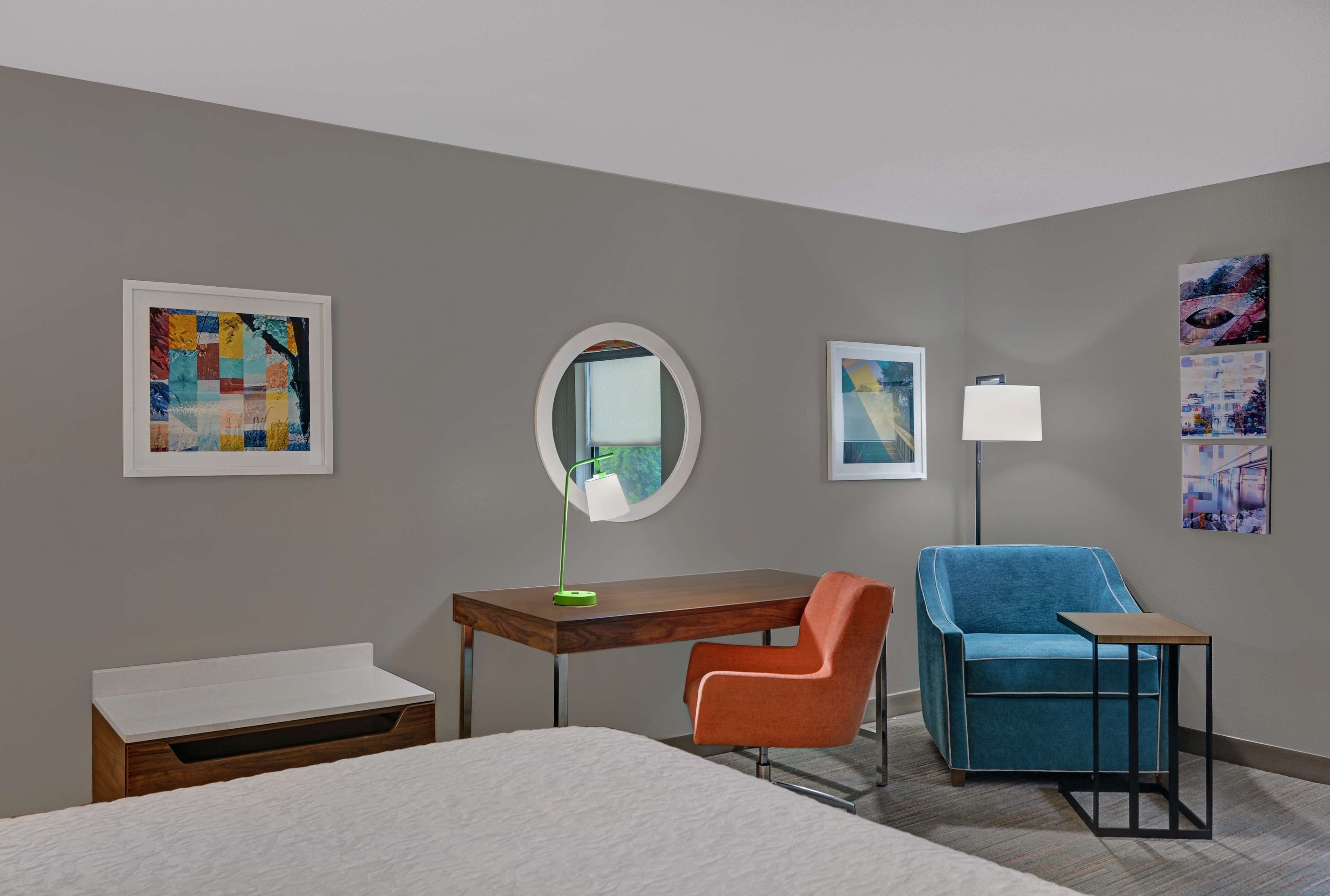 Una habitación de hotel moderna con una cama, un escritorio con lámpara, un sillón azul y obras de arte en las paredes.