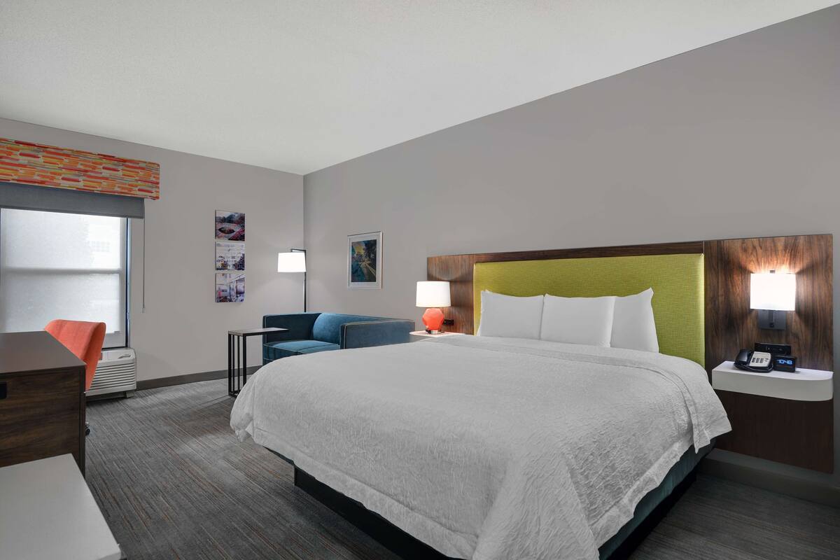 A hotel room with a large bed, a desk, a chair, a nightstand with a lamp, and a window with blinds.