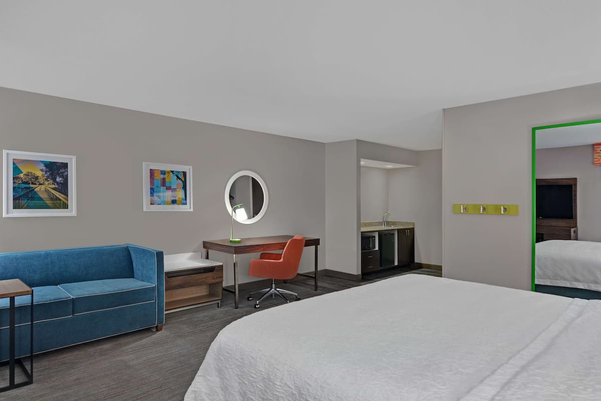 A hotel room with a bed, a desk with a chair, a sofa, a mirror, and artwork on the walls.