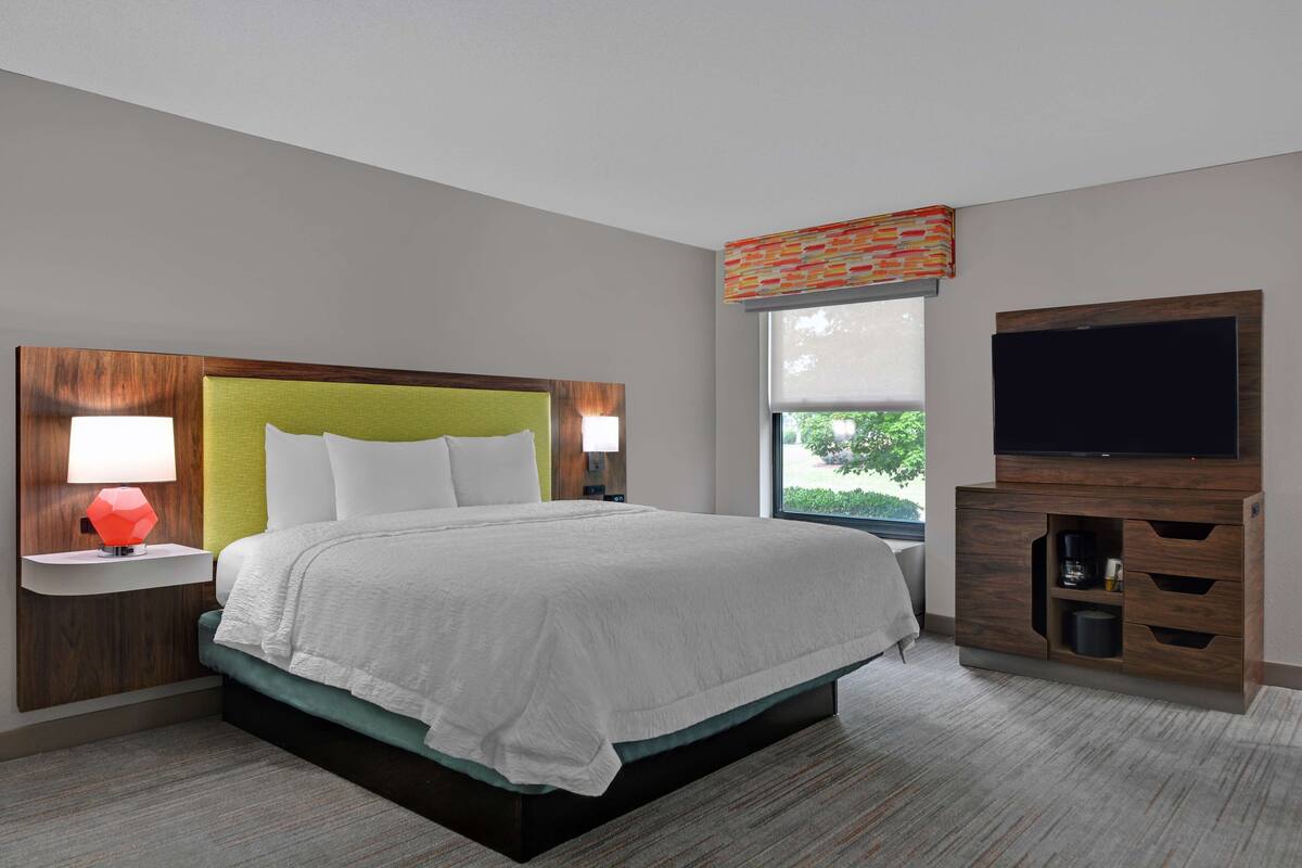 A modern hotel room with a large bed, a flat-screen TV, a nightstand with a lamp, and a window with a view of greenery.